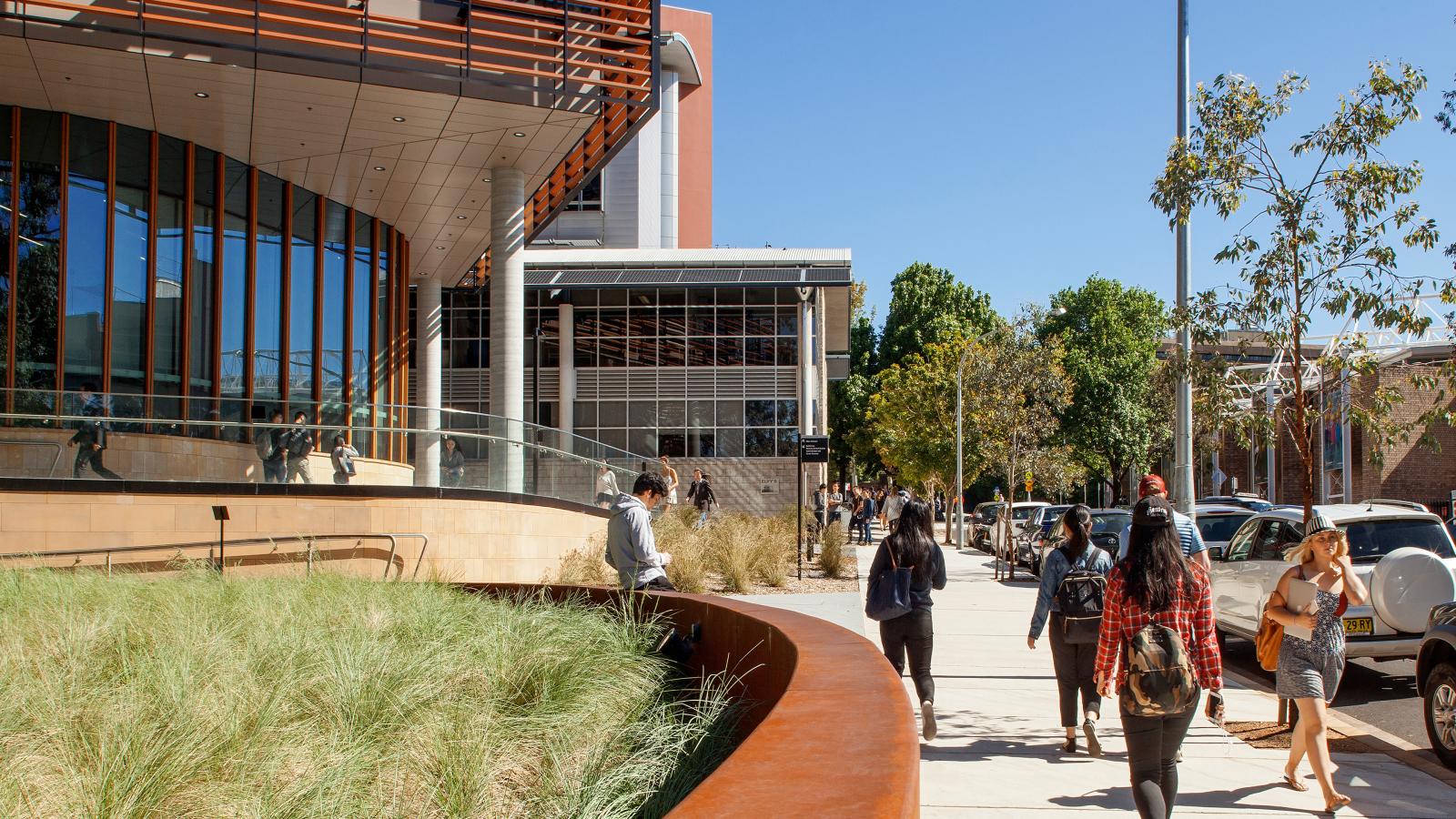 Students walk along a pathway filled with trees and grass on a sunny day, adjacent to the University of Sydney's modern Business School building with large glass windows and wooden accents. The background features more trees and additional buildings, creating a lively and studious atmosphere.