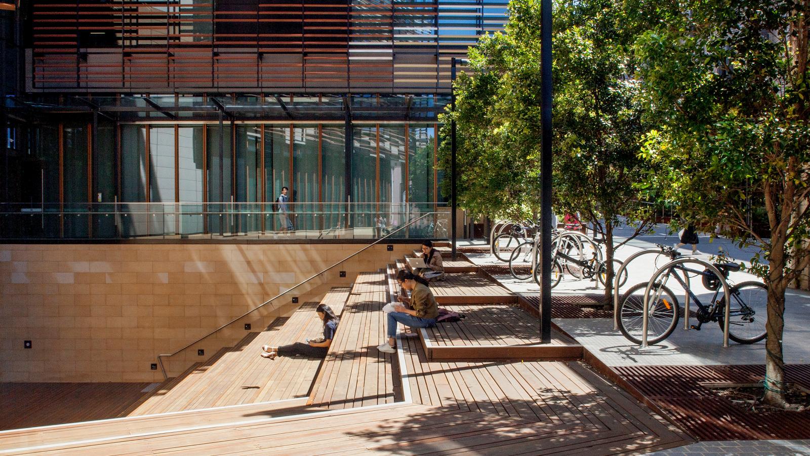 A modern outdoor seating area with wooden steps and platforms is shaded by trees. Bicycles are parked at racks to the right. A glass building with wooden slats in the background reflects sunlight. People are sitting and relaxing on the steps of the University of Sydney's Business School.