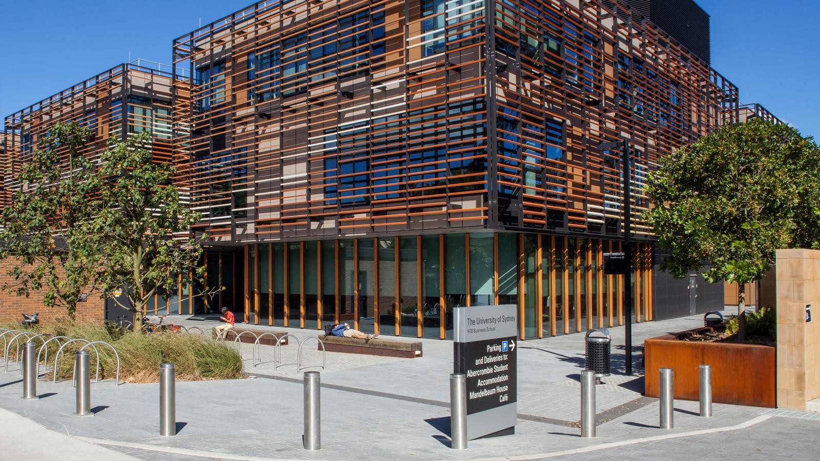 Modern building with a unique, structured facade made of horizontal and vertical beams. The Business School features large glass windows and is surrounded by a clean, paved area with bike racks, trees, and benches. A black sign with white text stands in front of the University of Sydney building.