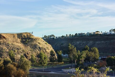 A serene landscape featuring a deep ravine with roads running along the bottom. The slopes of the ravine are dotted with trees and bushes. On the right, modern buildings and houses line the top of the ridge, offering a perfect lookout over the valley below. The sky above is clear with a few clouds.