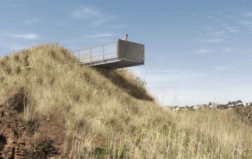 A person stands on a metal observation deck jutting out from a grassy hillside under a blue sky, overlooking a tranquil valley with sparse vegetation. To the right, buildings dot the distant landscape, offering a picturesque view from this scenic lookout.