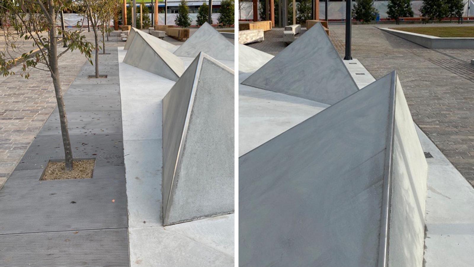 The image depicts several modern, triangular concrete sculptures arranged in Warrick Lane's public plaza. Each sculpture has a minimalistic design and is placed on a concrete base. Trees are planted along the edge of the plaza, with the surrounding area paved with stones.