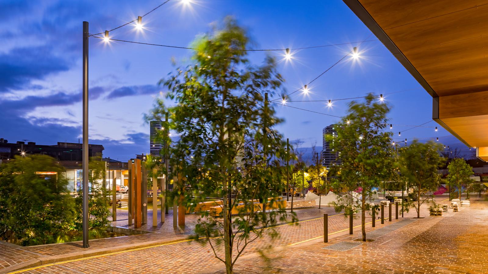 A well-lit urban street scene at dusk, showcasing string lights suspended above a walkway on Warrick Lane. Trees and benches line the path, adding a touch of nature. Modern buildings are visible in the background under a deep blue sky, hinting at a lively, vibrant atmosphere.