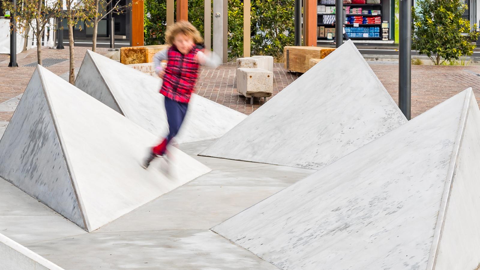 A child in a red and black plaid shirt and dark pants is in mid-air, jumping between large white concrete pyramid structures on Warrick Lane. In the background, there are benches, trees, and a building with a display window.