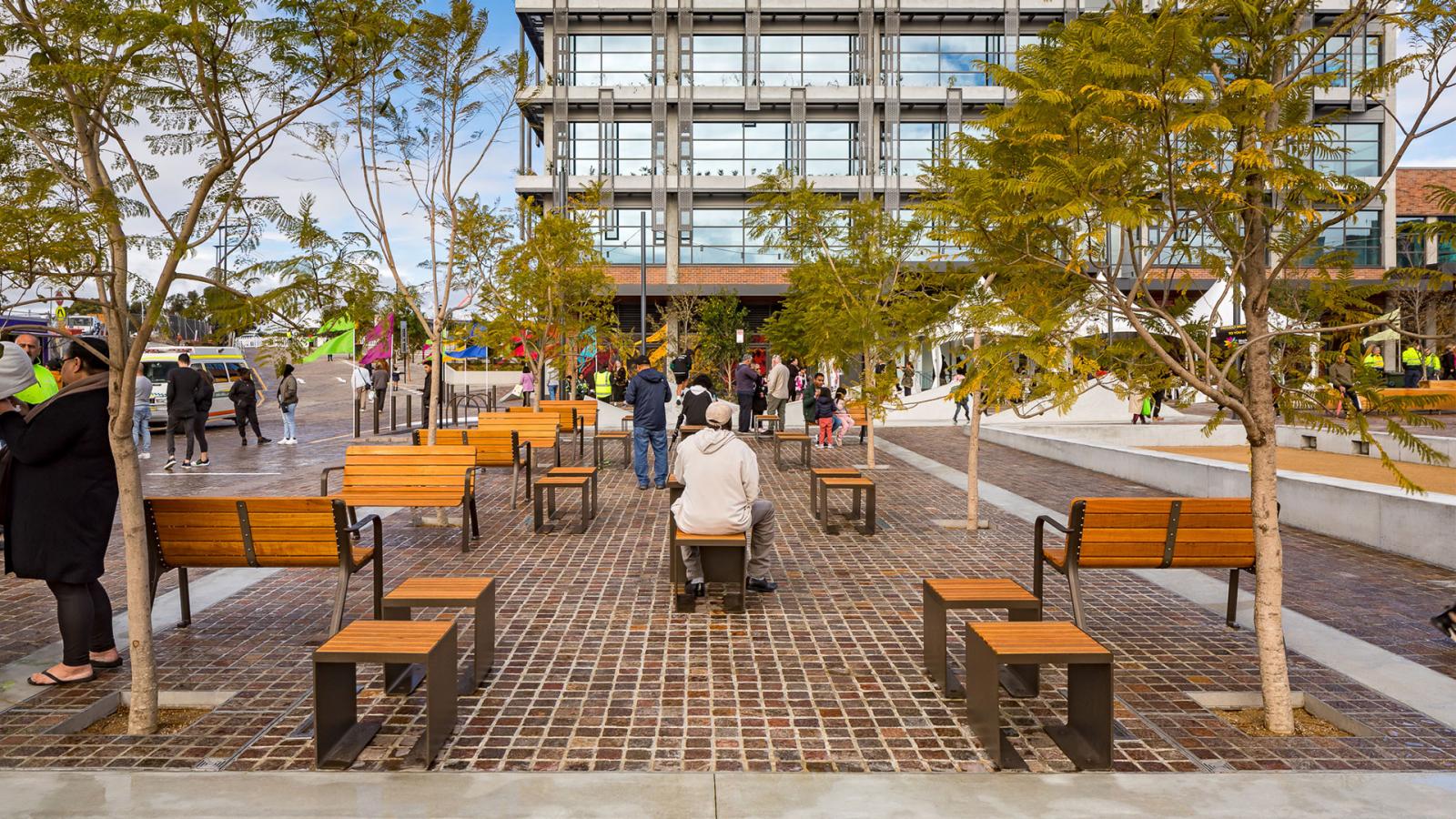 A modern urban plaza on Warrick Lane features several wooden benches and young trees on a paved surface. A person wearing a hat is seated in the center, looking at a group of people near a multi-story building with large windows. Some colorful decorations are visible in the background.