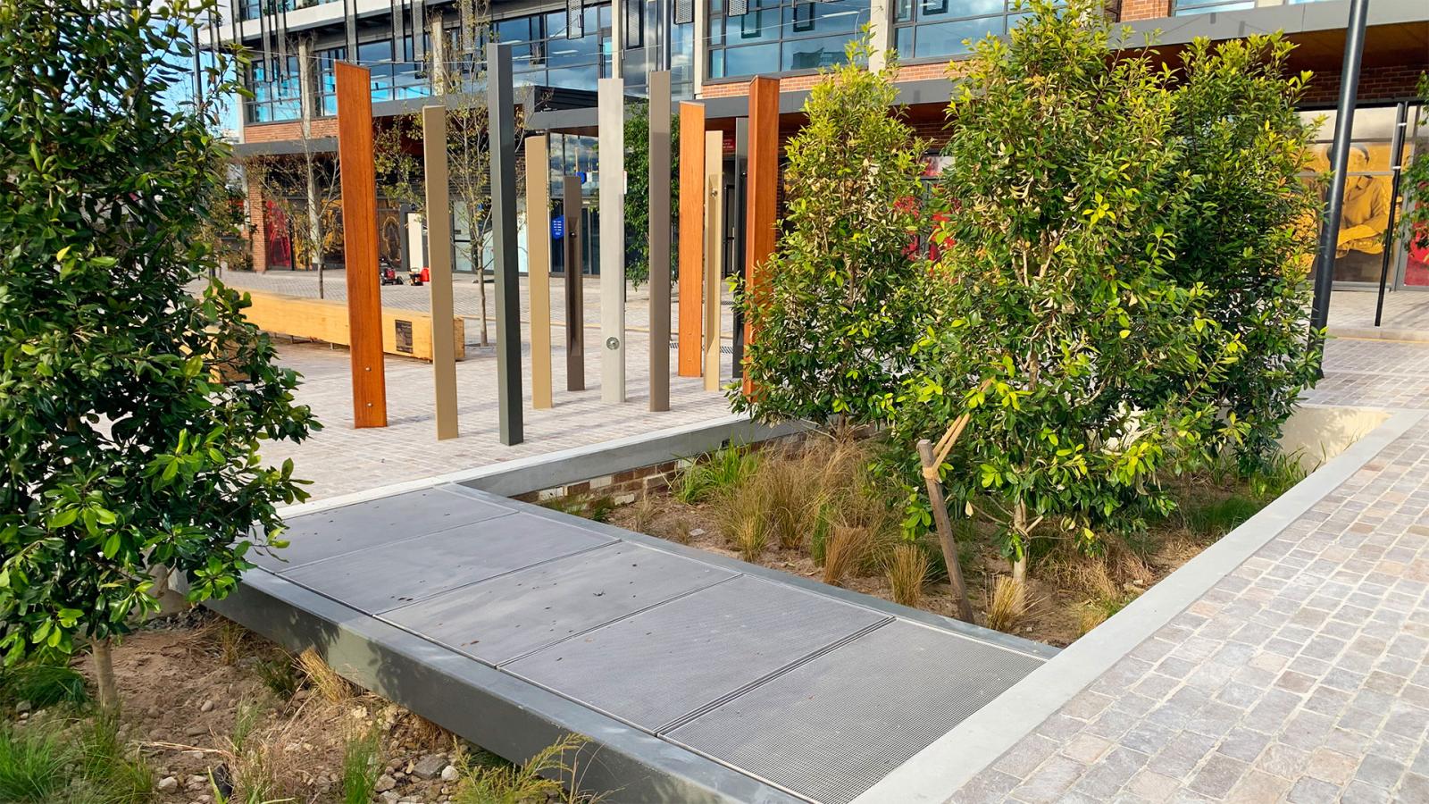 A modern outdoor walkway along Warrick Lane features metal and wooden vertical structures next to a building. The area boasts landscaped greenery, including young trees and grasses. The pathway is partially covered with metal panels and surrounded by a paved surface, adding an elegant touch.