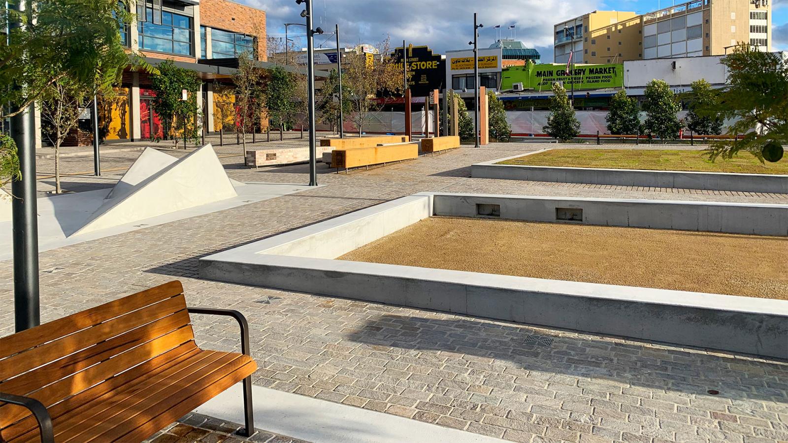 A modern urban plaza on Warrick Lane features concrete planters, brick walkways, wooden benches, and a mix of young trees. The background includes store signs and buildings under a partly cloudy sky. The area appears clean and minimalistic with a blend of natural and industrial elements.