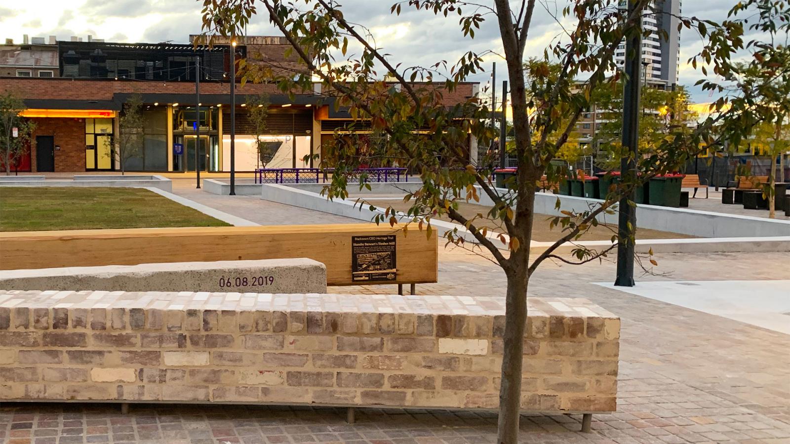 A modern outdoor plaza with stone benches and young trees along Warrick Lane. In the background are commercial buildings with large glass windows, and there is a mix of paved and grassy areas. The date "06.08.2019" is engraved on one of the stone benches.