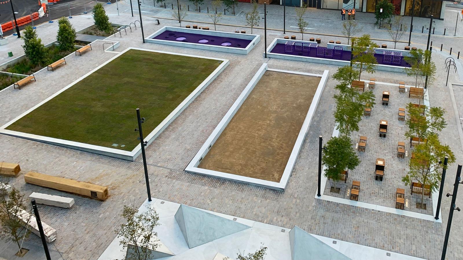 Aerial view of Warrick Lane's large, modern, open public space featuring three rectangular areas: one with grass, another with sand, and the third with purple seating. Adjacent to them are rows of wooden benches, tables with chairs, young trees, and paved walkways.