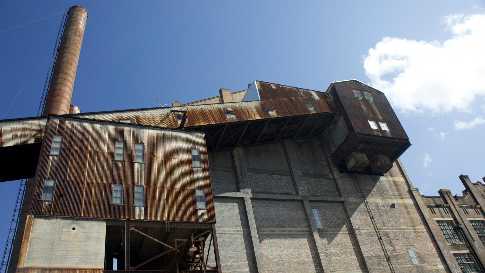 A low-angle view of an old industrial building with rusted metal siding and a tall chimney at White Bay Power Station. The structure exudes a weathered appearance with multiple windows and protruding sections, showcasing its historic urban design framework against a clear sky with a few clouds.