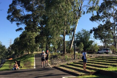 People are enjoying an outdoor park scene along the Yarra River. One person appears to be taking photos, while others are walking and exercising along a paved path surrounded by lush trees and greenery, highlighting the area's rich biodiversity with another car parked and the blue sky visible in the background.