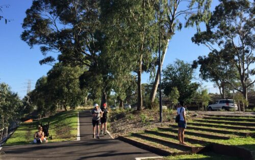 People are enjoying an outdoor park scene along the Yarra River. One person appears to be taking photos, while others are walking and exercising along a paved path surrounded by lush trees and greenery, highlighting the area's rich biodiversity with another car parked and the blue sky visible in the background.