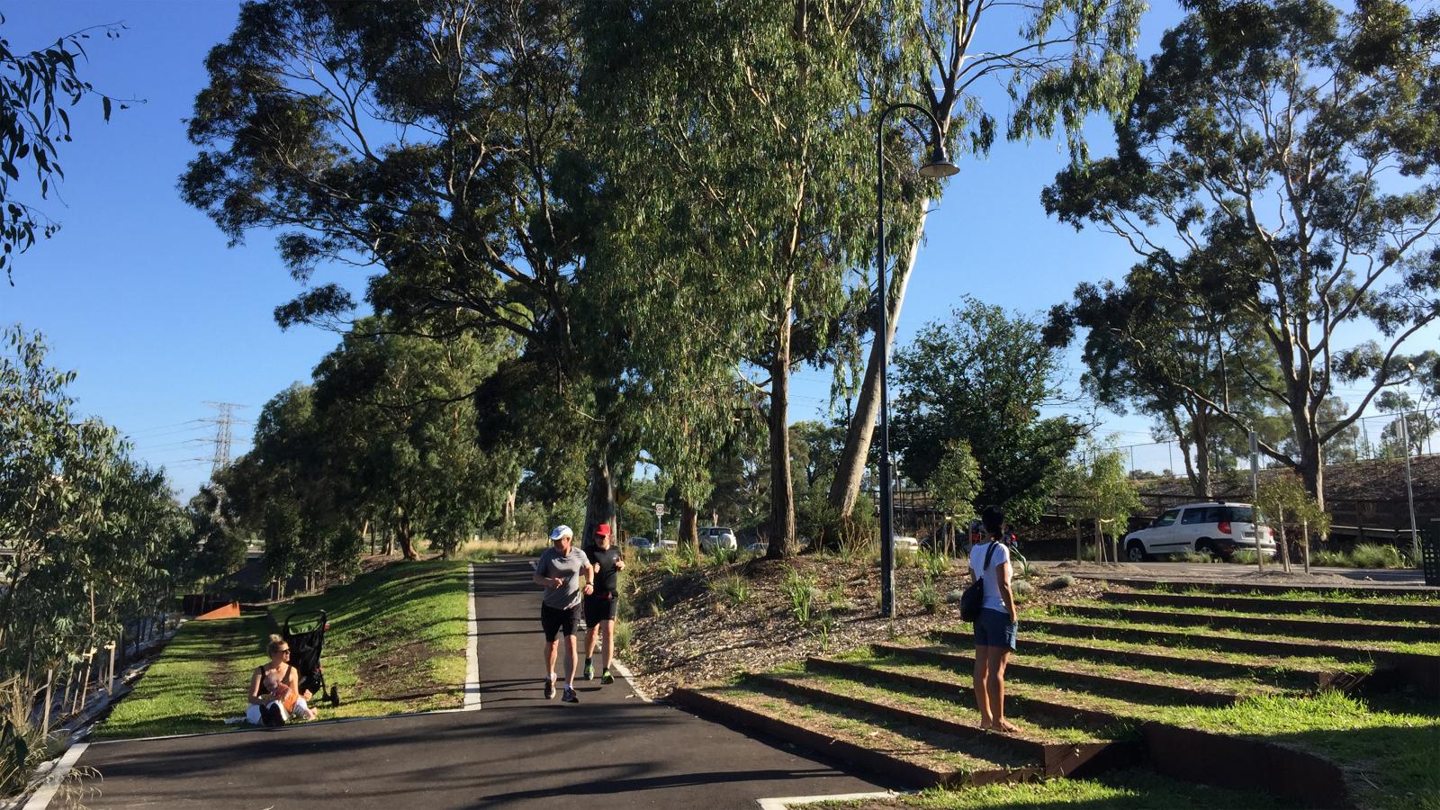 People are enjoying an outdoor park scene along the Yarra River. One person appears to be taking photos, while others are walking and exercising along a paved path surrounded by lush trees and greenery, highlighting the area's rich biodiversity with another car parked and the blue sky visible in the background.