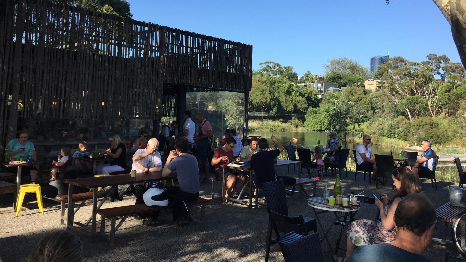 A bustling outdoor café with people seated at various tables enjoying drinks and conversation, all set near the Yarra River. Surrounded by lush greenery, a modern building with a wood-slatted facade is visible on the left. It's a sunny day with a clear blue sky, enhancing the area's rich biodiversity.