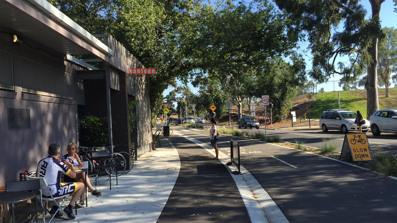 A sunny street with cyclists and pedestrians. Two people sit outside a building with a "Kanteen" sign, near bicycles. A cyclist rides on a marked bike lane beside a pedestrian path linked to the Yarra River. Trees line the road, and a "SLOW DOWN" sign is next to the bike lane.