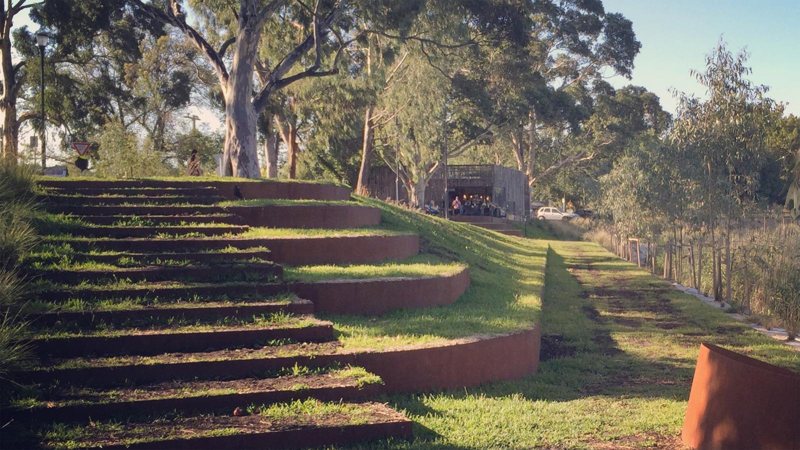 A grassy amphitheater with several ascending rows of seating leads to a wooden structure in the background. Trees surround the area, creating a natural and serene environment that enhances local biodiversity. People are visible near the wooden structure, suggesting a gathering or event.