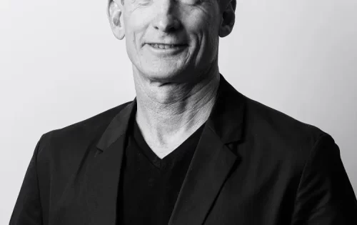 Black and white portrait of Adrian McGregor, a middle-aged man with short, curly hair wearing a dark blazer over a V-neck shirt. He is facing the camera and smiling slightly against a plain background.