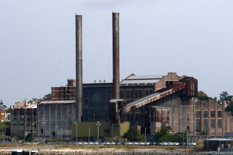 A large, abandoned industrial building resembling the White Bay Power Station stands near a body of water, featuring two tall smokestacks and several rusted structures. The sky above is overcast, and numerous parked cars are visible in front of the building. Sparse vegetation surrounds the area.