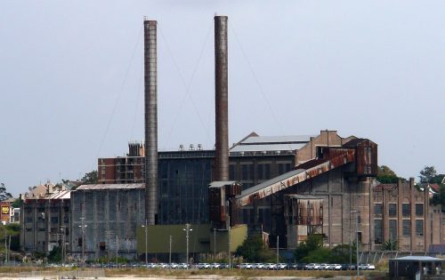 A large, abandoned industrial building resembling the White Bay Power Station stands near a body of water, featuring two tall smokestacks and several rusted structures. The sky above is overcast, and numerous parked cars are visible in front of the building. Sparse vegetation surrounds the area.