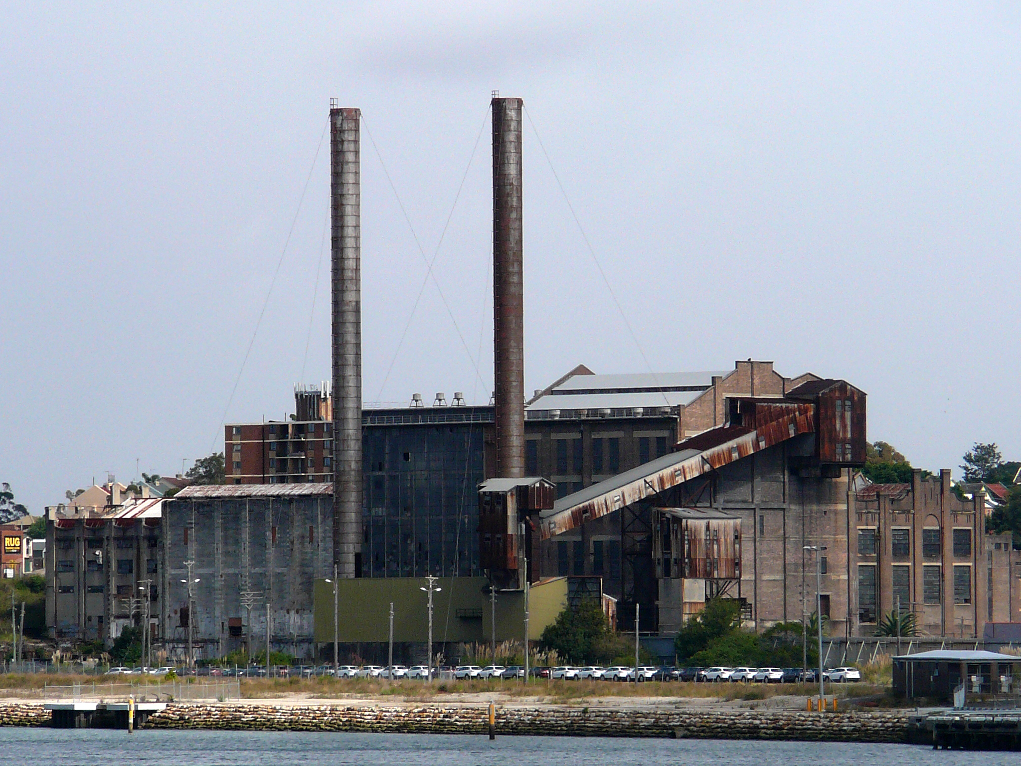 A large, abandoned industrial building resembling the White Bay Power Station stands near a body of water, featuring two tall smokestacks and several rusted structures. The sky above is overcast, and numerous parked cars are visible in front of the building. Sparse vegetation surrounds the area.