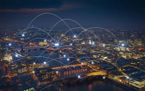 Aerial view of a cityscape at night, with illuminated buildings and bridges over a river. White light trails arc over the City of London, connecting different points, symbolizing digital connectivity for smarter cities. The sky is dark, with a hint of twilight on the horizon.