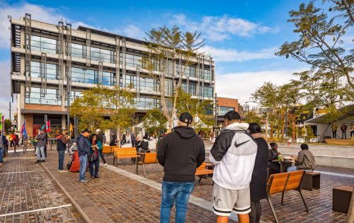 A lively outdoor public space with people walking and sitting on benches along Warrick Lane. The area is paved with cobblestones and has several trees planted along walkways. In the background, there's a modern multi-story building with large windows and a clear blue sky overhead.