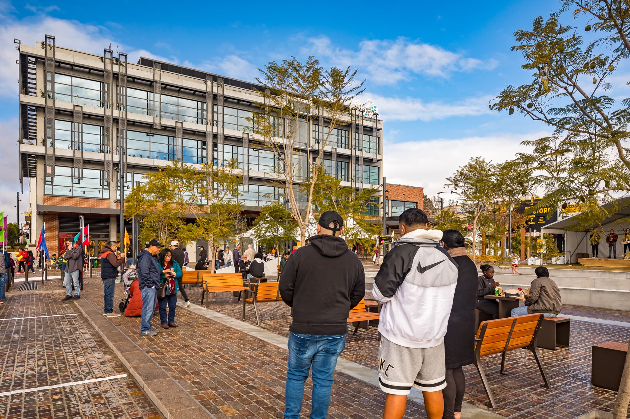 A lively outdoor public space with people walking and sitting on benches along Warrick Lane. The area is paved with cobblestones and has several trees planted along walkways. In the background, there's a modern multi-story building with large windows and a clear blue sky overhead.