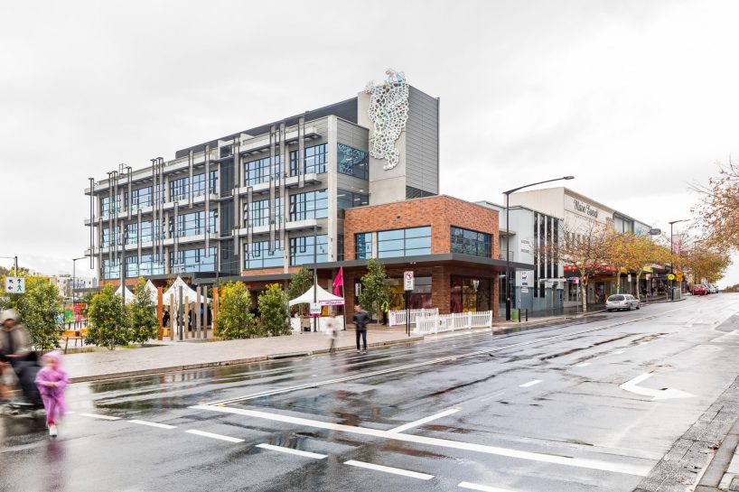 A modern multi-story building with glass windows and decorative artwork on its facade stands on Warrick Lane, a wet street. A small park with structures and greenery is next to it. People are walking, some with umbrellas, under a cloudy sky after rain.