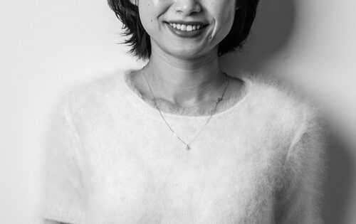 Black and white portrait of Ann Deng, a woman with short dark hair smiling at the camera. She is wearing a short-sleeved, fluffy light-colored sweater and a delicate necklace with a small pendant. She stands against a plain background.