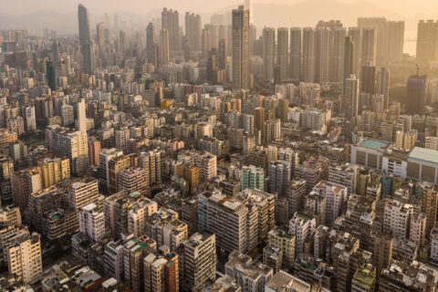 Aerial view of a densely packed urban area with many high-rise buildings. The cityscape, demonstrating elements of biourbanism, is surrounded by mountains in the background under a hazy sky at sunset. The landscape shows a mix of residential and commercial buildings integrated with green spaces.