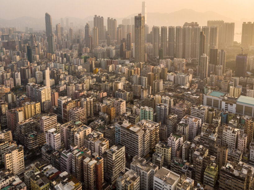 Aerial view of a densely packed urban area with many high-rise buildings. The cityscape, demonstrating elements of biourbanism, is surrounded by mountains in the background under a hazy sky at sunset. The landscape shows a mix of residential and commercial buildings integrated with green spaces.