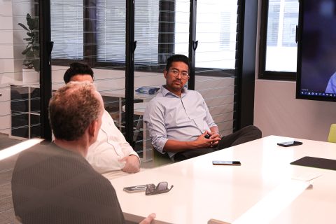 Three men are sitting around a conference table in a modern office. One man faces the other two, with Dr. Yazid Ninsalam listening intently, a phone and notebook in front of him. There is a large screen in the room, possibly for presentations or video calls.