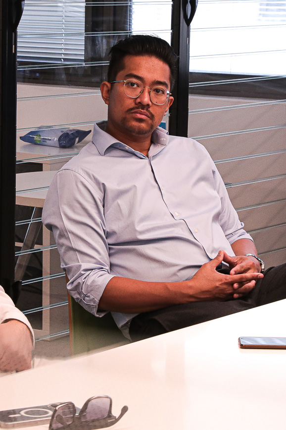 Dr. Yazid Ninsalam, a man with short hair and glasses, wearing a light blue shirt and dark pants, sits at a table with his hands clasped. The background shows part of an office with a computer keyboard and glasses visible on the table.