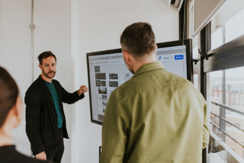 Michael Cowdy, dressed in a black jacket, points at a large screen displaying various images while talking to two colleagues, one of whom is wearing a green jacket. They are in a bright room with large windows.