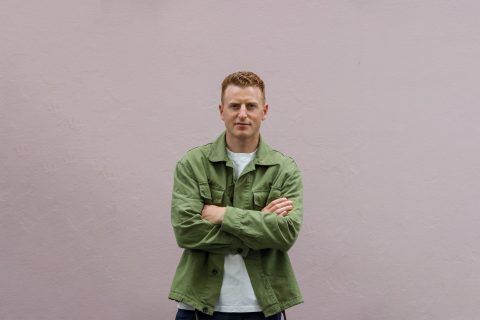 Man stands against a plain wall with his arms crossed, wearing a green jacket over a white shirt. Fraser Halliday's short hair is styled neatly, and he has a calm expression as he looks directly at the camera. The background is a simple, light-colored textured wall.