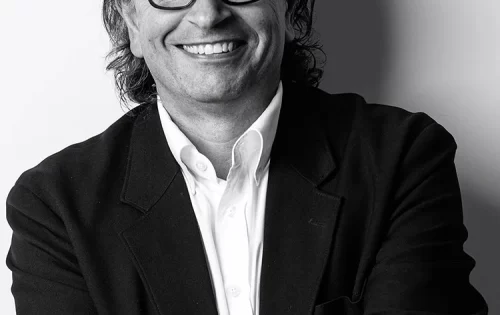 Black-and-white photo of George Panos, a smiling man with dark, wavy hair wearing glasses, a white dress shirt, and a dark blazer. He stands against a plain background, arms crossed, displaying a watch on his left wrist.