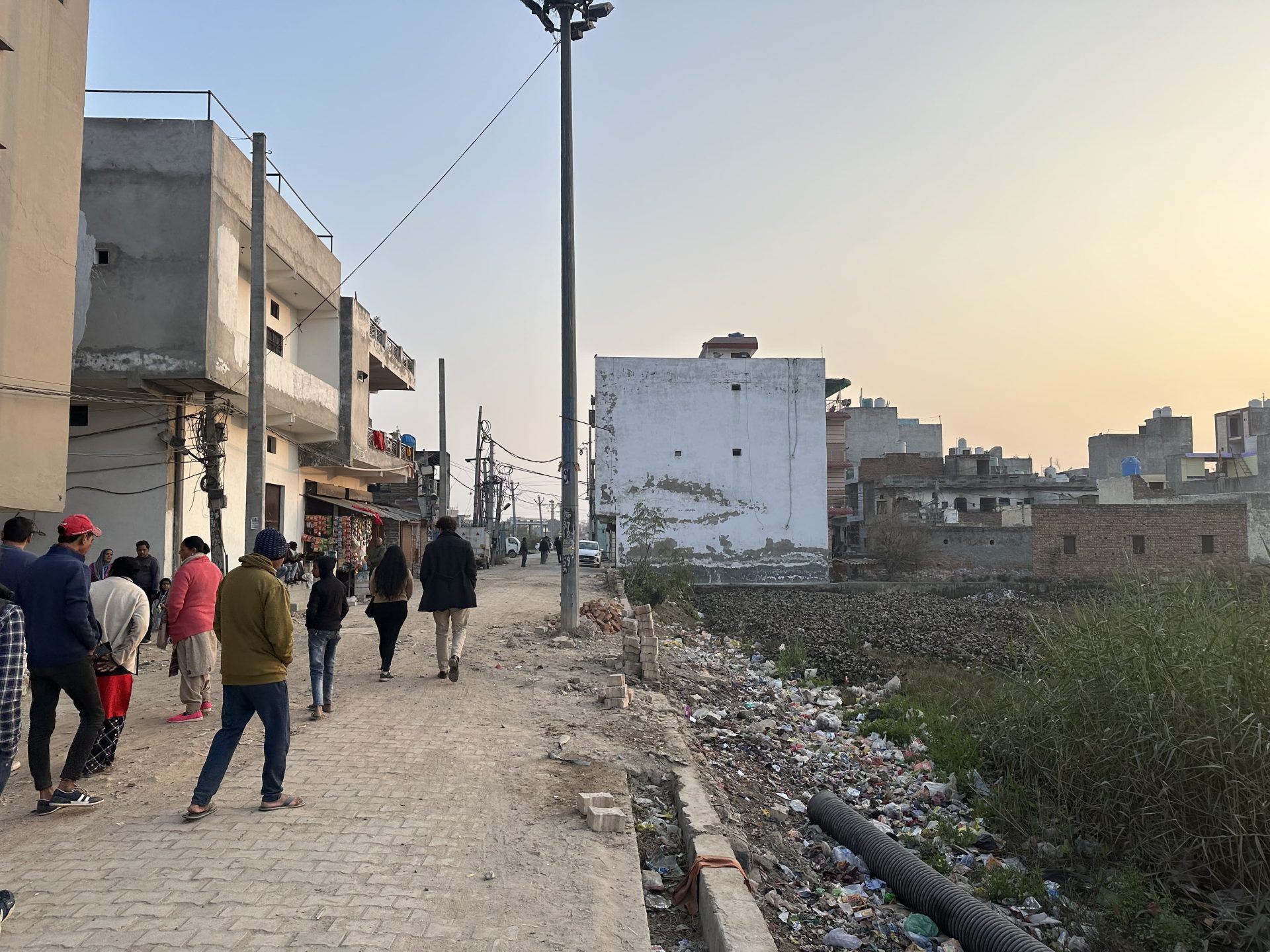 A group of people walks along a pathway beside a canal or drainage area in an urban neighborhood. Buildings are visible on both sides, some unfinished or under construction. Electrical wires hang overhead, and the sky is clear with a hint of sunset, highlighting the efforts of the Australia India Water Security Initiative (AIWASI).