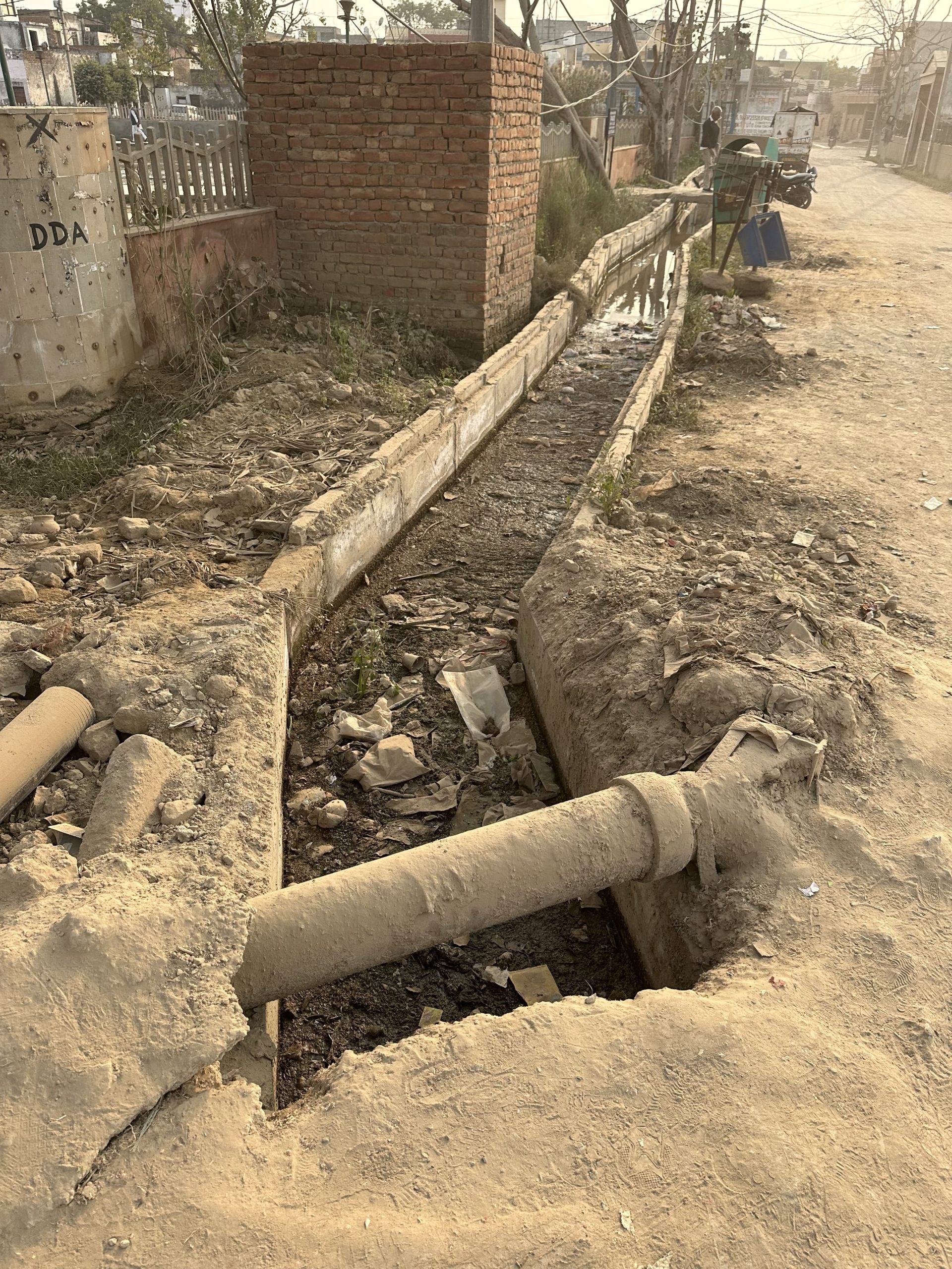 An open trench with exposed pipes, part of the AIWASI Water Initiative, runs alongside a road in an underdeveloped area. Dirt and debris surround the trench, and a brick structure stands nearby. A few vehicles and trees are visible in the background along the dusty road.