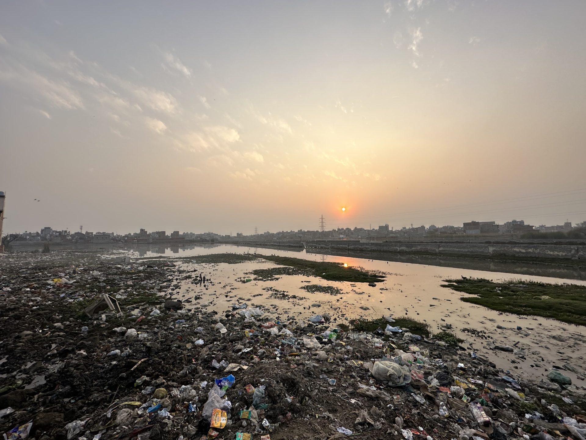 A polluted riverbank cluttered with trash and debris, including plastic and metal waste. The sky is hazy with clouds, illuminated by the setting sun. Buildings and power lines are visible in the distant background. This scene of environmental neglect underscores the urgency for initiatives like AIWASI, aimed at improving water security for Australia and India.