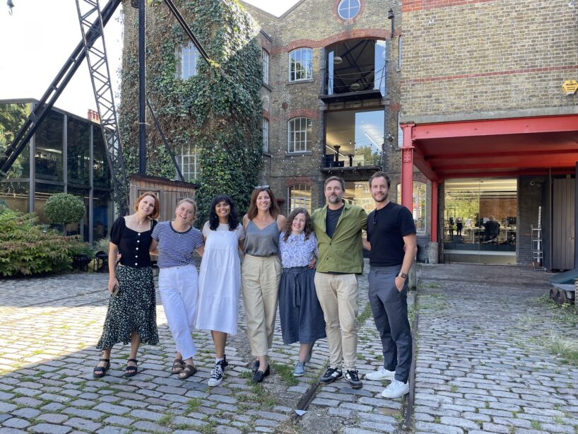 A group of seven people stand closely together, posing for a photo in a cobblestone courtyard in front of London Studio's brick building covered in ivy. Everyone is smiling, and they are dressed casually on a sunny day.