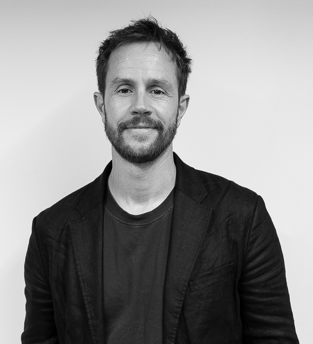 Black and white photo of Michael Cowdy with short hair and a beard, smiling slightly, wearing a dark blazer over a plain T-shirt, standing in front of a plain background.