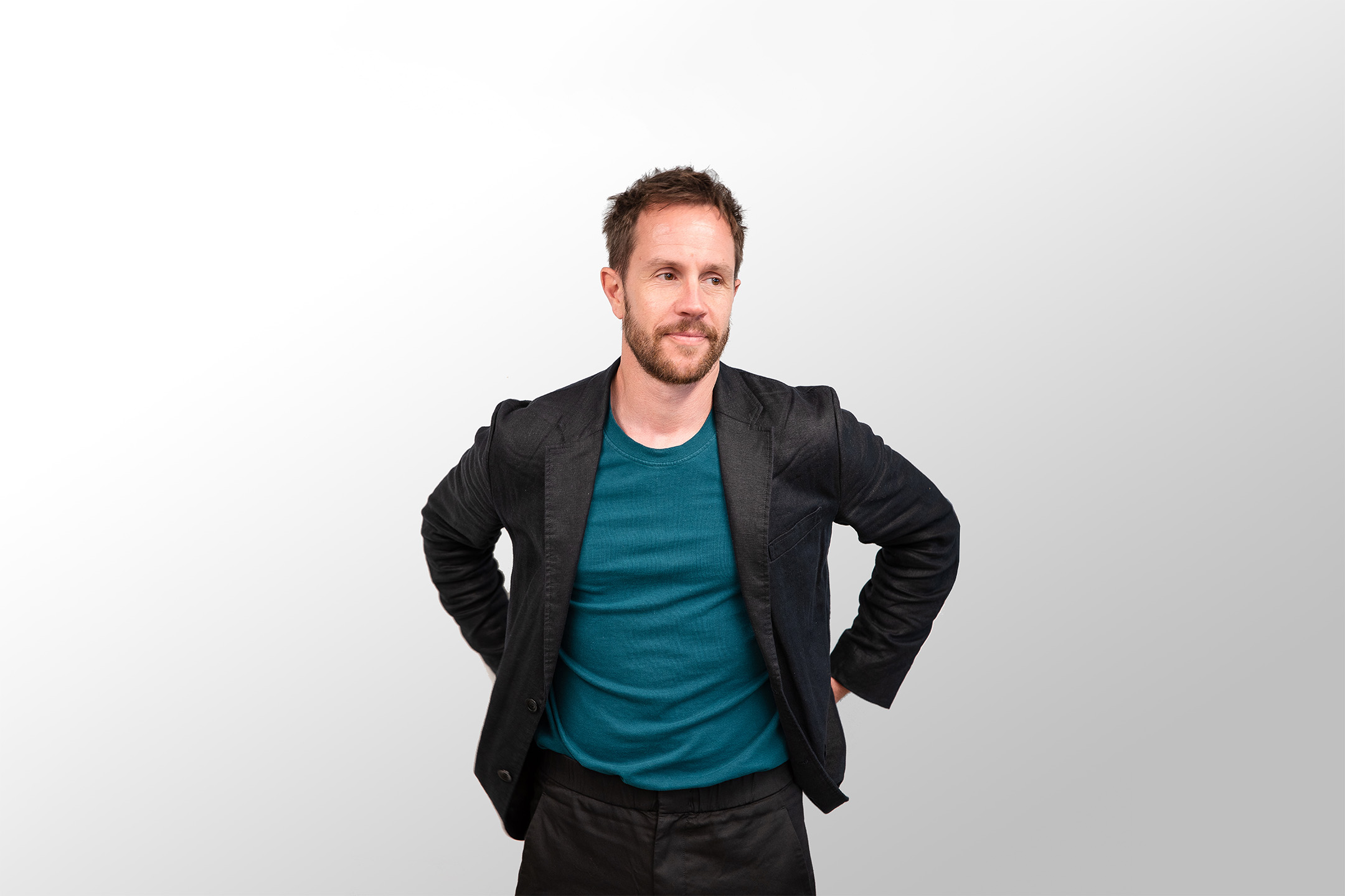 Michael, a man with short hair and a beard, stands against a plain white background. He is wearing a dark blazer over a teal shirt and has his hands resting on his hips. He appears to be looking slightly off-camera, exuding an air of casual confidence.