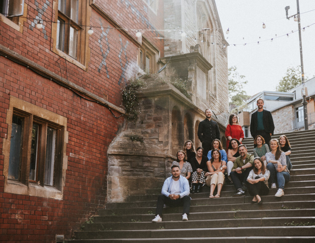 A group of 15 people are sitting and standing on outdoor stairs next to a brick and stone building in Bristol. String lights hang above them, with greenery visible in the background. Everyone is smiling and facing the camera, capturing a delightful studio-like shot in this charming locale.