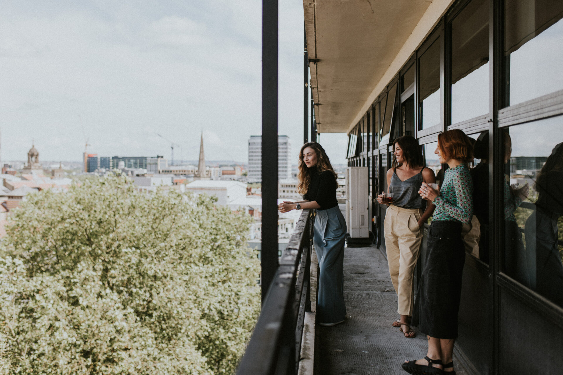 Three women are standing on a balcony overlooking the Bristol cityscape with trees and buildings in the background. One woman is leaning on the railing, while the other two are engaged in conversation. The setting appears to be a bright, sunny day.