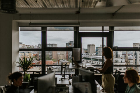 A modern office space with large windows offering a cityscape view. Three people are working at their desks with computer monitors. Natural light illuminates the room, and a potted plant is placed near a window. The atmosphere appears busy yet focused, embodying the essence of our "About Us" story.