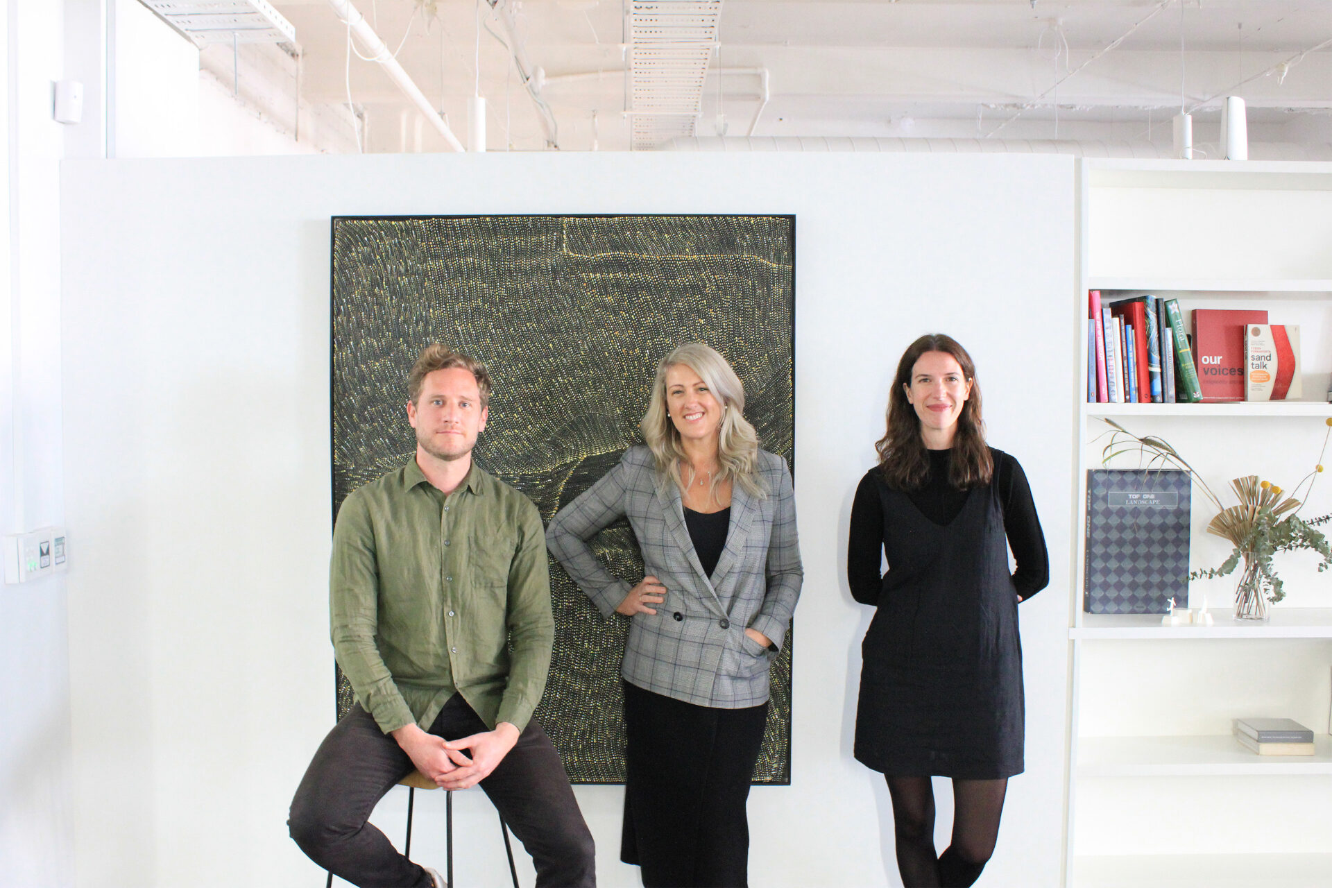 Three people are posing indoors in the McGregor Coxall Melbourne studio in front of a black and gold artwork. The person on the left, Nick Griffin, sits on a stool wearing a green shirt, the person in the middle, Katie Earle, stands with a hand on their hip in a gray plaid blazer, and the person on the right, Miranda Wilkinson, stands in a black dress.