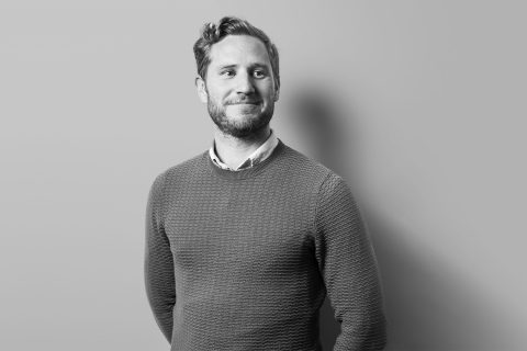 Black and white photo of Nick Griffin standing against a plain wall. He has short hair, a beard, and is smiling slightly. He is wearing a textured sweater over a collared shirt and has his hands behind his back.