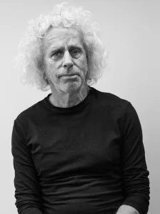 Black-and-white portrait of Philip Coxall, an older man with curly, white hair and a beard. He is wearing a long-sleeved, black shirt and is looking directly at the camera with a neutral expression. The background is plain and light colored.