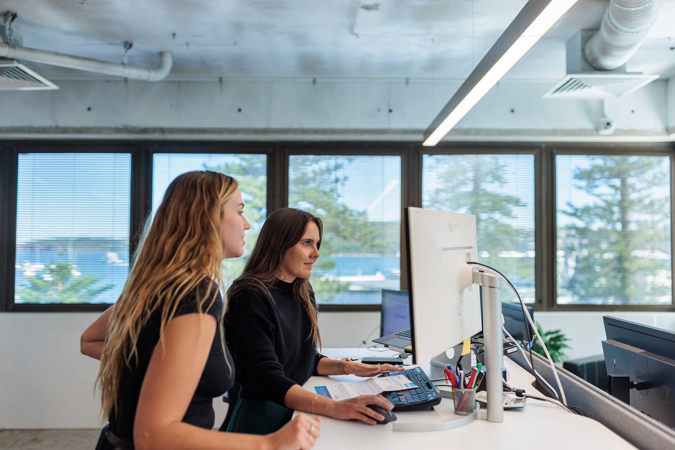 Two women are working together at a desk in a modern Manly office with large windows providing a view of trees, Sydney Harbour and blue sky outside. One woman is seated and typing on a keyboard, while the other stands beside her, both focused on a computer screen in their collaborative studio space.