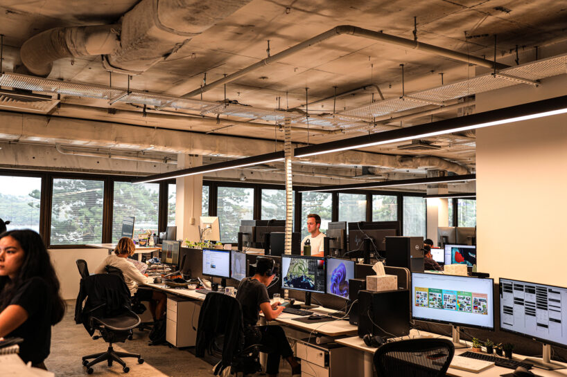 Modern office space with exposed ceilings, large windows, and multiple workstations. Office workers are seated at desks with computer monitors, while one person stands. Outside the windows, a hilly Sydney landscape with greenery is visible.